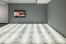 Innenansicht einer Kunstgalerie mit minimalistischer Einrichtung, einem auffälligen, bunten Kunstwerk an der Wand und einem einzigartigen, leuchtenden Boden.